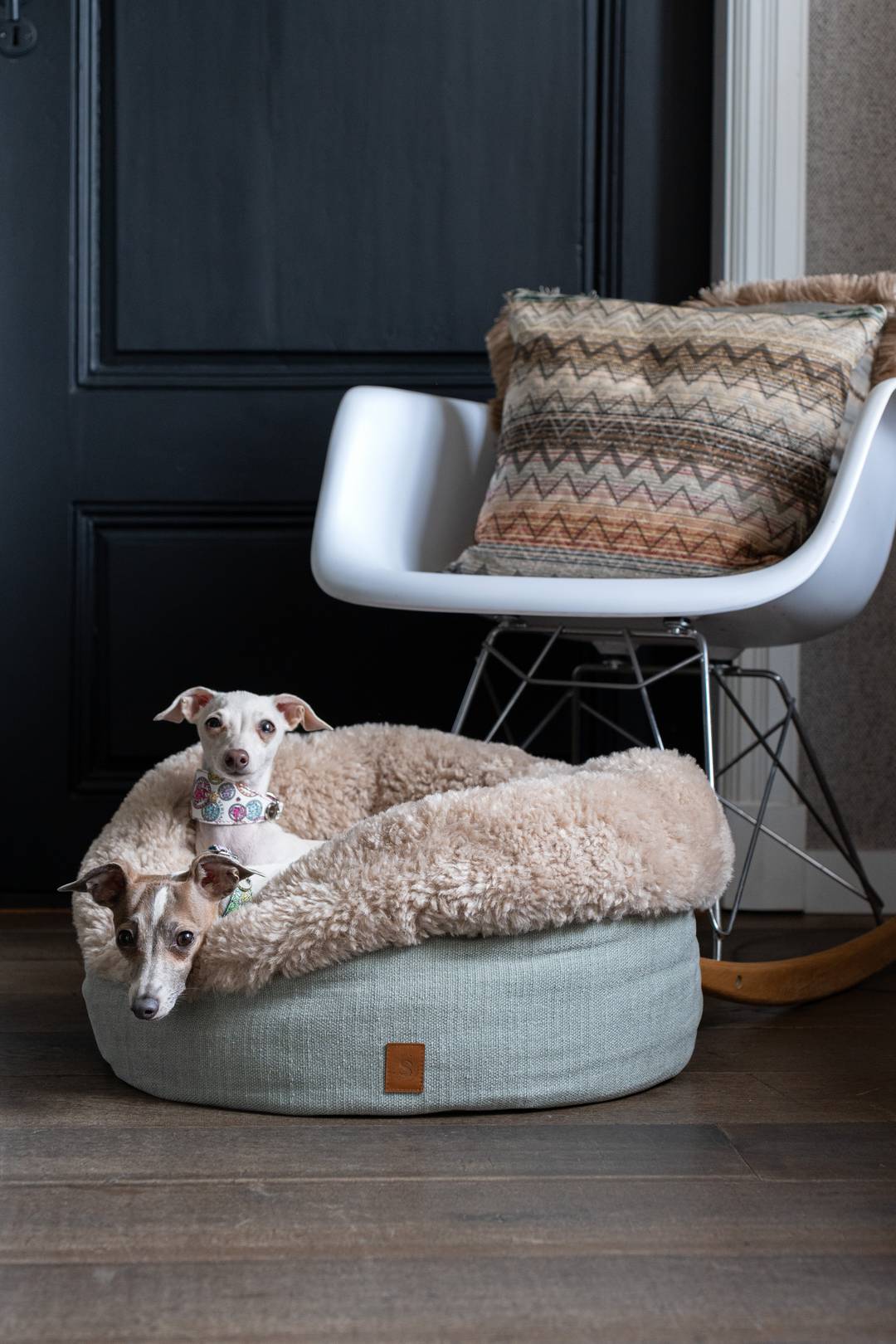 Stijlvolle hondenmand met 2 whippets erin geplaatst bij een witte stoel en zwarte deur.